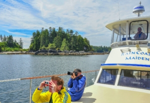 Front of tour boat with Captain at helm & 2 male guests sitting on bow looking at island through binoculars and camera